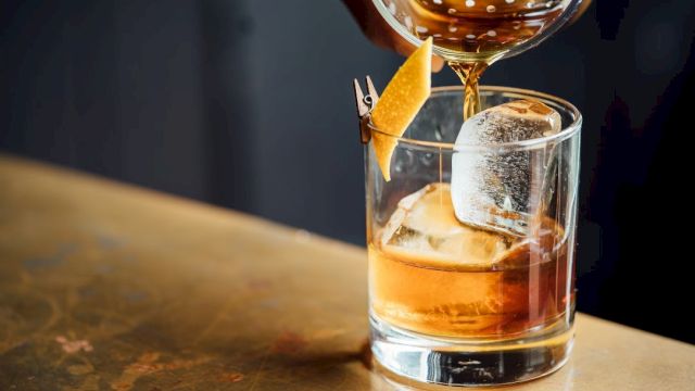 A whiskey glass with large ice cube, an orange twist, and a drink being poured from a bottle over a wooden bar top.