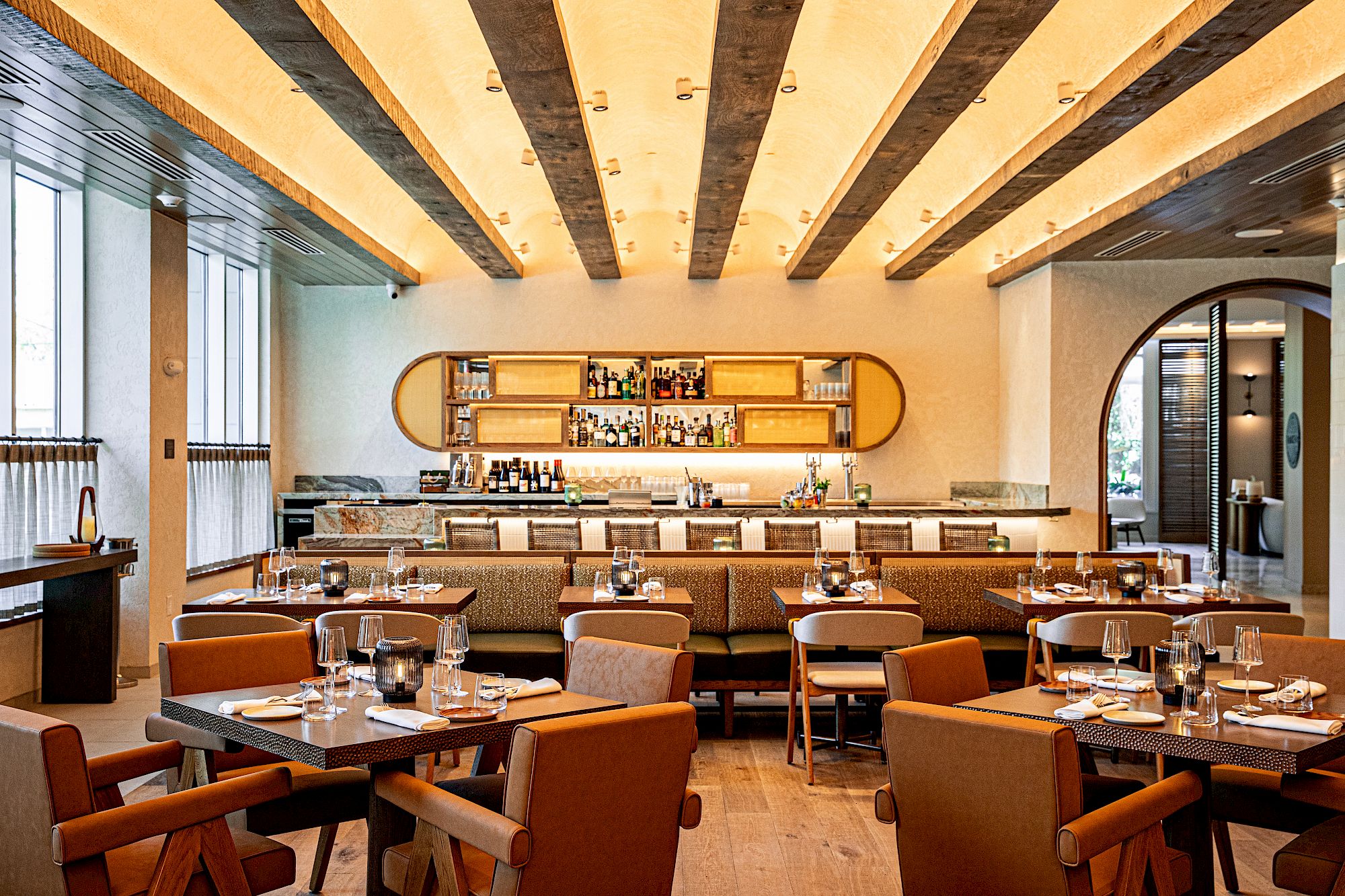 A stylish dining room with wooden beams, warm lighting, and a long shelf displaying items; tables set with glassware and neatly arranged chairs.