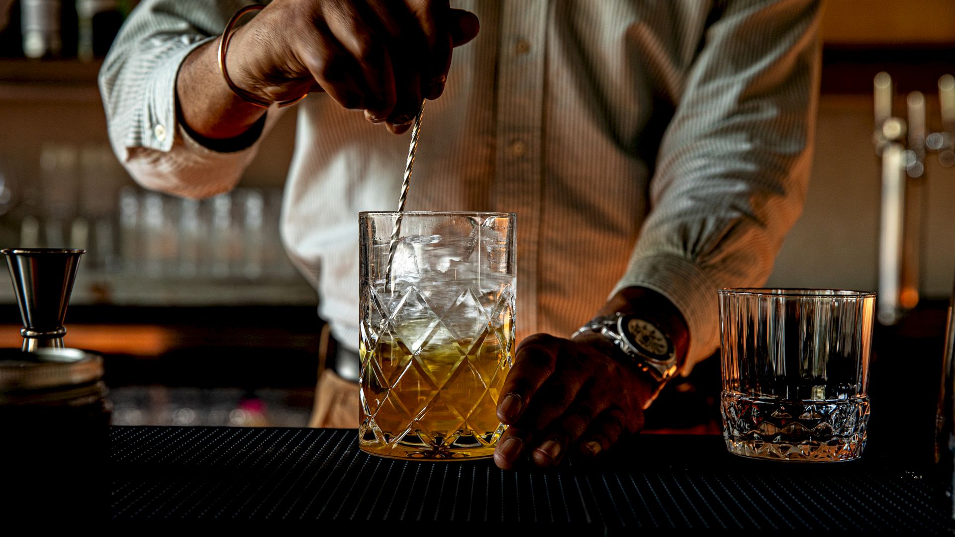 A bartender stirs a drink with a twist of ice, glassware on the bar, and a moody, dim-lit atmosphere.
