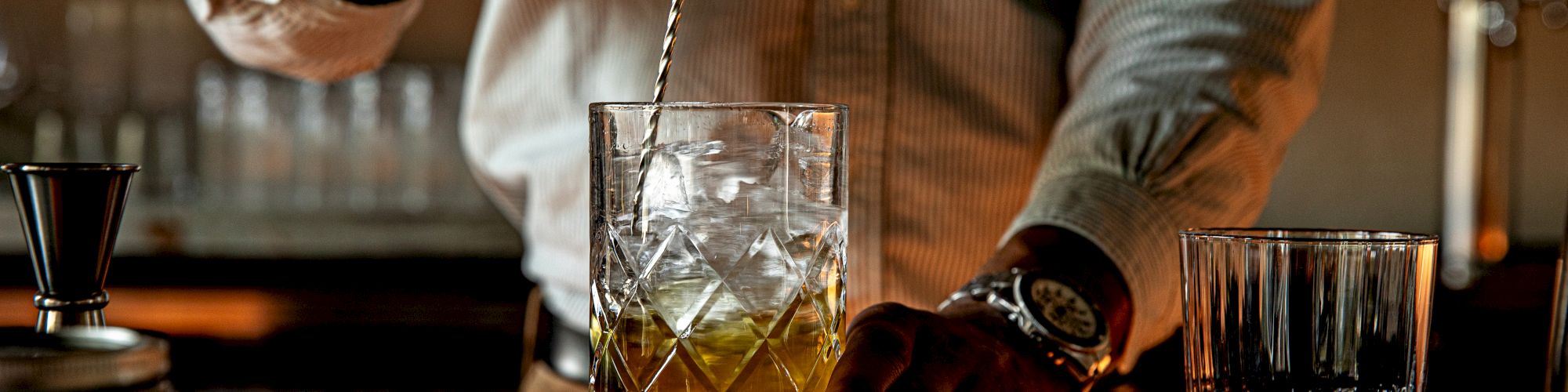 A bartender stirs a drink with a twist of ice, glassware on the bar, and a moody, dim-lit atmosphere.