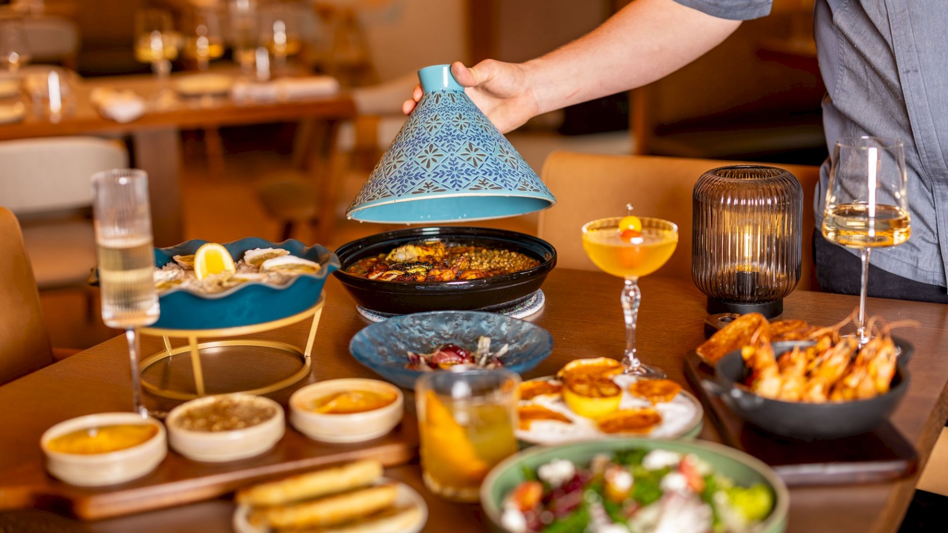 A colorful table of shared dishes with dips, grilled skewers, salads, bread, and drinks; a person lifts a blue cloche over a pot, inviting feast.