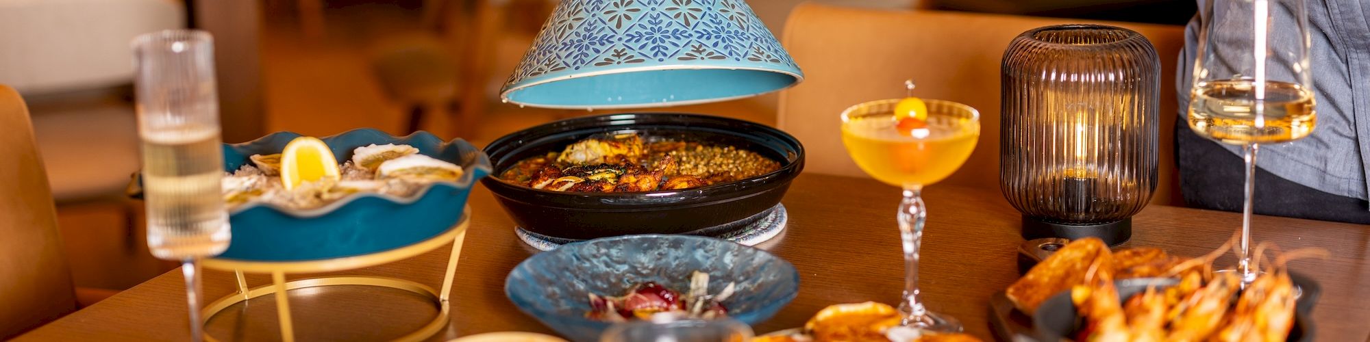A colorful table of shared dishes with dips, grilled skewers, salads, bread, and drinks; a person lifts a blue cloche over a pot, inviting feast.
