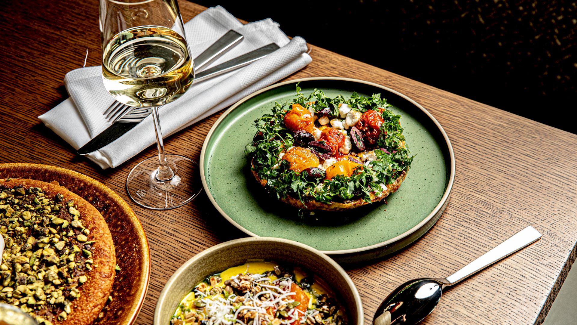 A glass of white wine with a pastry, a kale-topped plate, and a colorful grain bowl on a wooden table with utensils nearby.