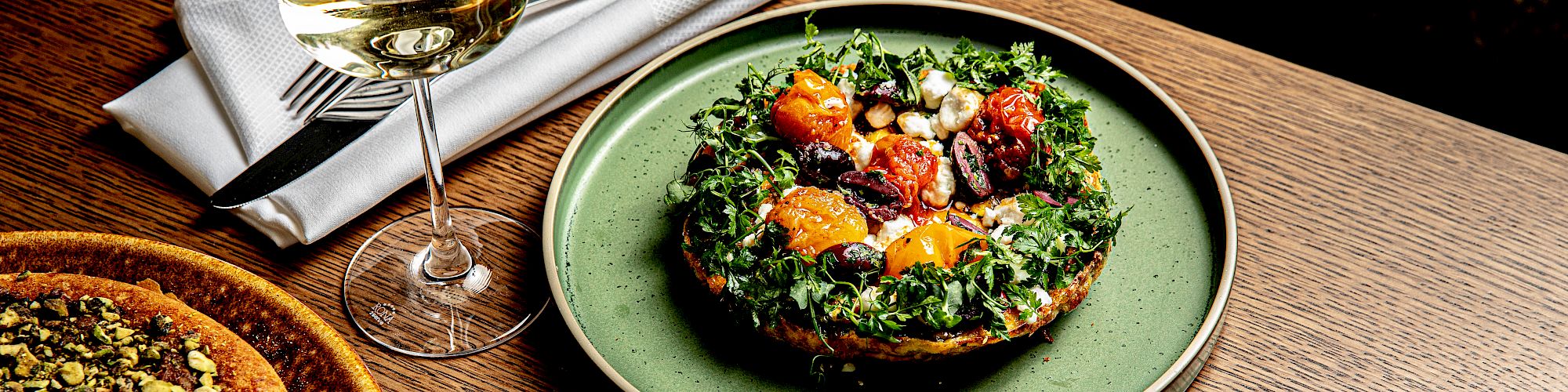 A glass of white wine with a pastry, a kale-topped plate, and a colorful grain bowl on a wooden table with utensils nearby.