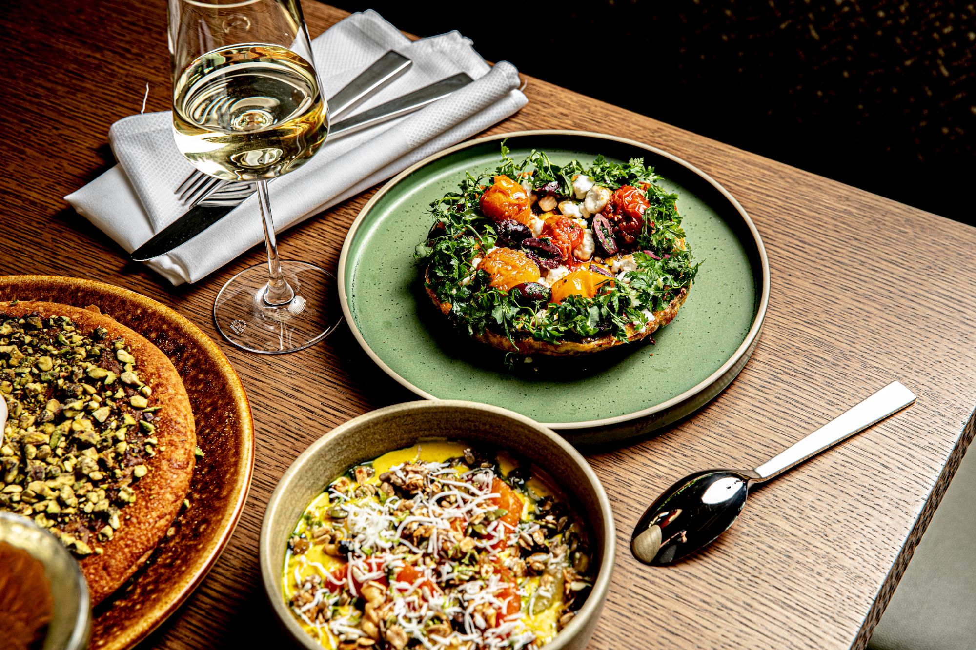 A glass of white wine beside a table with two plates of colorful food: a green plate with a kale and toppings dish, and a brown bowl with a mixed salad, plus a spoon and napkins.