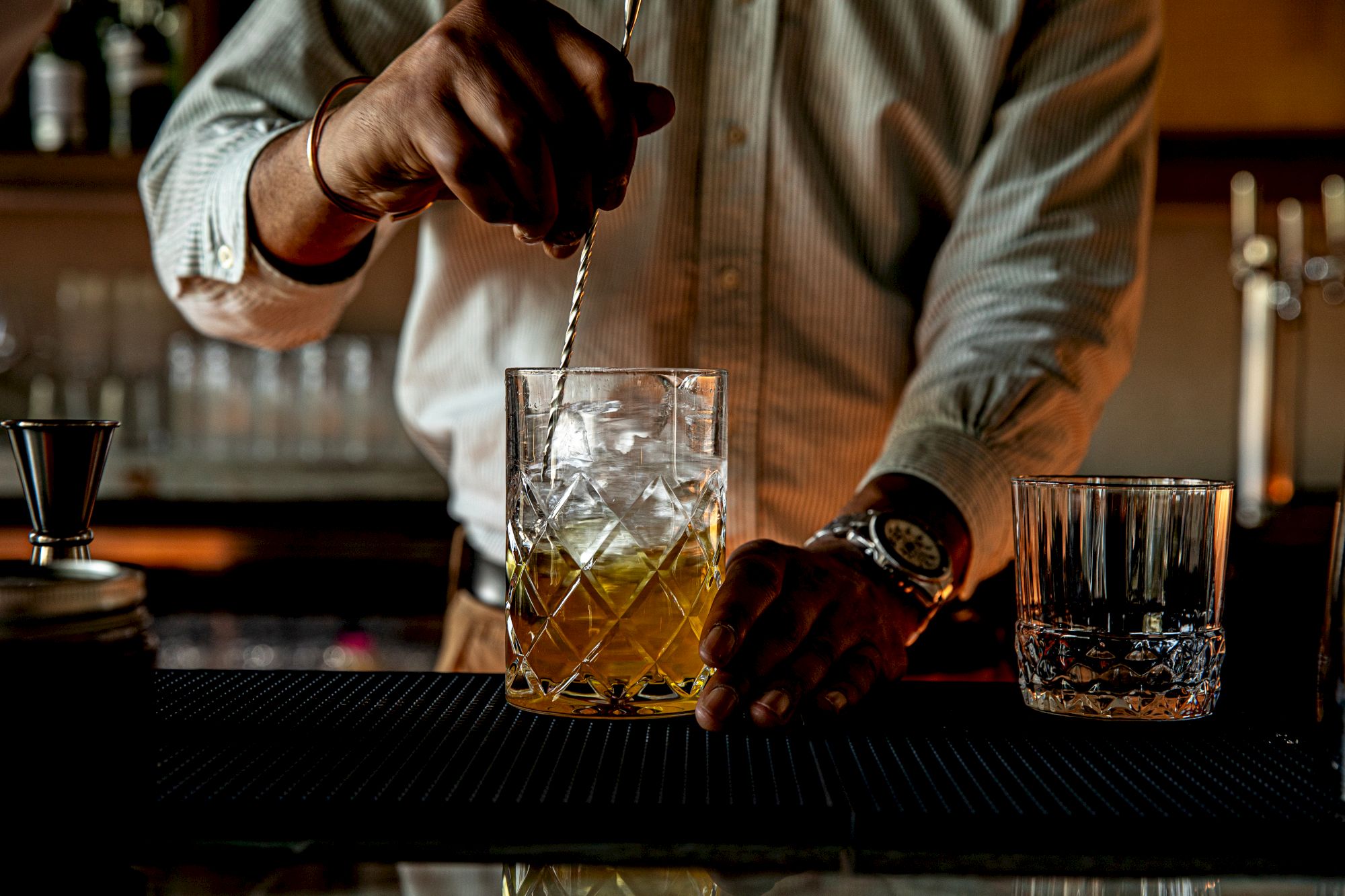 A bartender stirs a drink in a crystal glass with ice, bar tools, and a rocky pour over a dark bar counter.