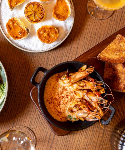 A top-down meal spread: salad bowl, sliced citrus on a plate, shrimp bake in a small pot, toasted bread, and drinks on a wooden table, delicious.