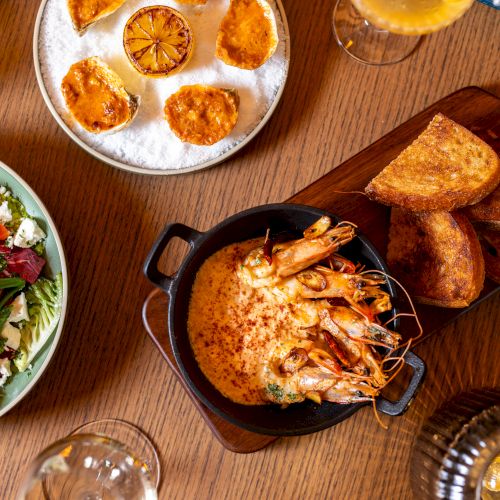 A top-down meal spread: salad bowl, sliced citrus on a plate, shrimp bake in a small pot, toasted bread, and drinks on a wooden table, delicious.