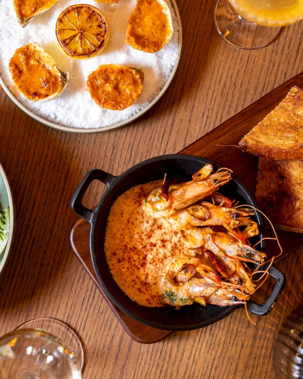 A top-down meal spread: salad bowl, sliced citrus on a plate, shrimp bake in a small pot, toasted bread, and drinks on a wooden table, delicious.