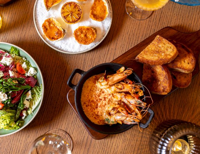 A top-down meal spread: salad bowl, sliced citrus on a plate, shrimp bake in a small pot, toasted bread, and drinks on a wooden table, delicious.