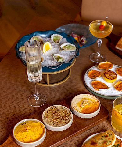 A table with seafood oysters on a blue platter, several dips and sauces, bread with herbs, and drinks including a champagne flute and a cocktail, all arranged invitingly.