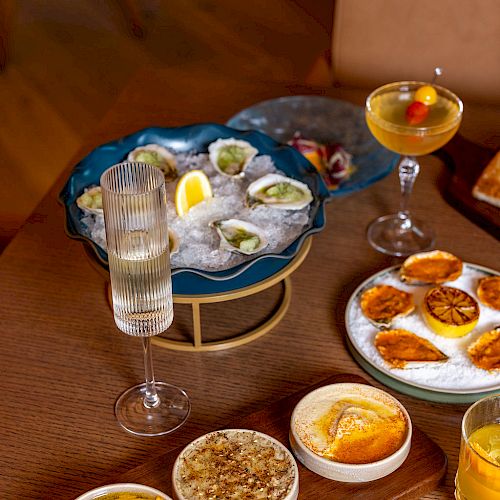 A table with seafood oysters on a blue platter, several dips and sauces, bread with herbs, and drinks including a champagne flute and a cocktail, all arranged invitingly.