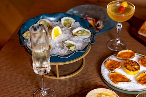 A table with seafood oysters on a blue platter, several dips and sauces, bread with herbs, and drinks including a champagne flute and a cocktail, all arranged invitingly.