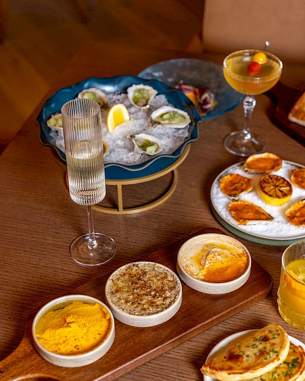A table with seafood oysters on a blue platter, several dips and sauces, bread with herbs, and drinks including a champagne flute and a cocktail, all arranged invitingly.