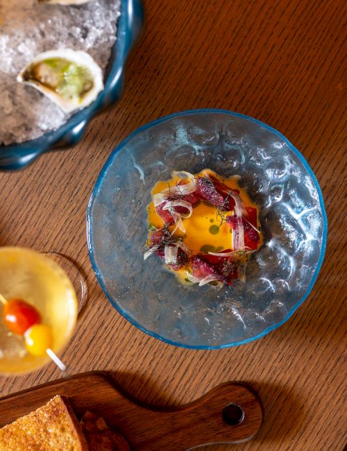 A blue glass bowl with a fried egg and colorful toppings sits on a wooden table, alongside a plate of tacos and a cork-board cutting board.