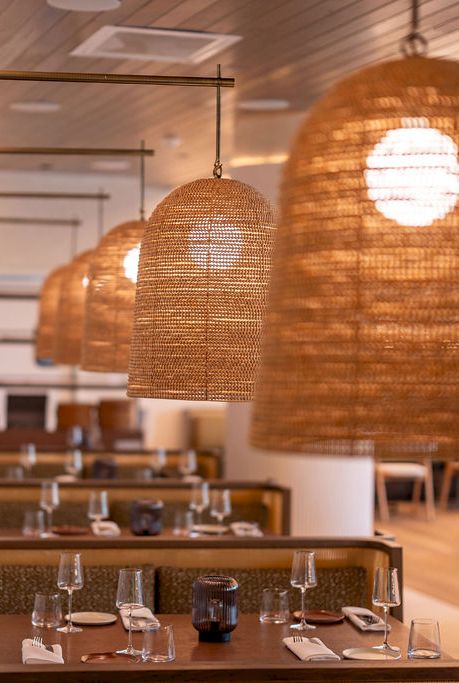 A cozy restaurant interior with woven pendant lamps, wood tables, and neatly set place settings along a softly lit dining area.