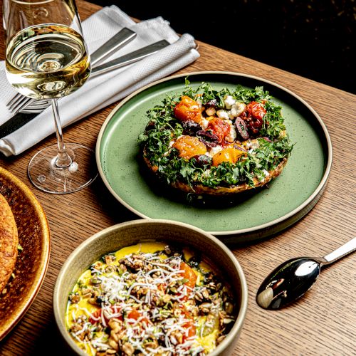 A wooden table with a wine glass, a green plate of salad, a bowls of colorful grain bowls, and a slice of nut-topped cake, ready to eat.