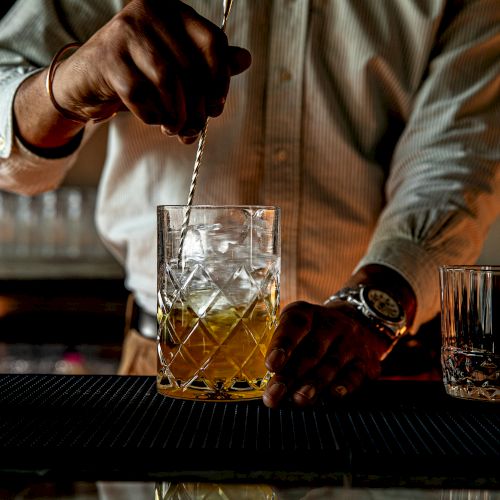 A bartender stirs a whiskey cocktail in a glass with ice, on a bar counter, while a separate glass sits nearby, creating a moody bar scene.