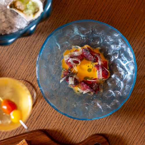 A colorful, half-melted dessert sits in a blue glass bowl with toppings like fruit and cream, on a wooden table with bread nearby.