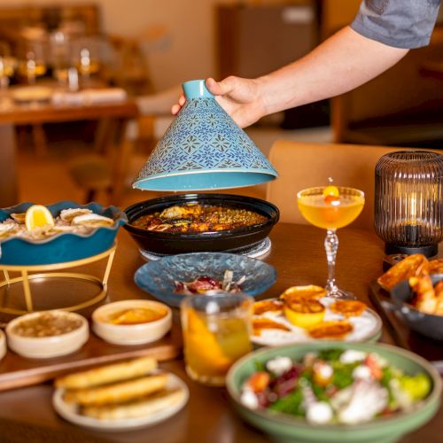A colorful feast on a table with tapas, dips, bread, grilled veggies, cocktails, and a pot of steaming stew being stirred under a blue chimney.