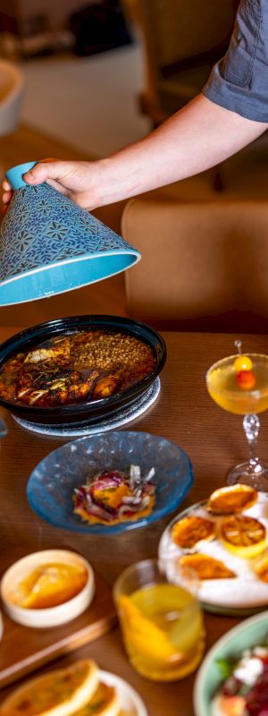 A person lifts a blue cloche over a spread of tapas and dishes on a dining table, with candles and wine, in a warm ambient setting.