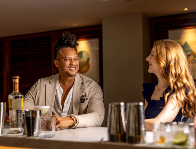 Two people sit at a table on a panel or discussion stage, with drinks and papers in front of them, talking. Ends with a period.