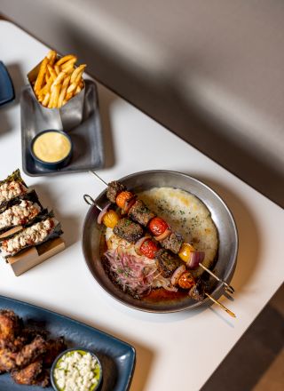 A skillet with sizzling meat and veggies, a skewer of grilled sausages, and creamyDip or sauce nearby, forming a tasty barbecue spread.