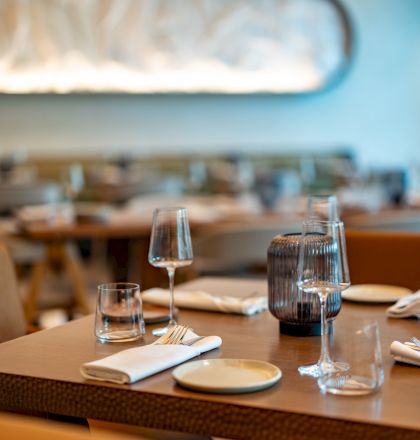 A dining table set with plates, glasses, and cutlery in a cozy restaurant, blurred background, warm lighting, and a glass of water.