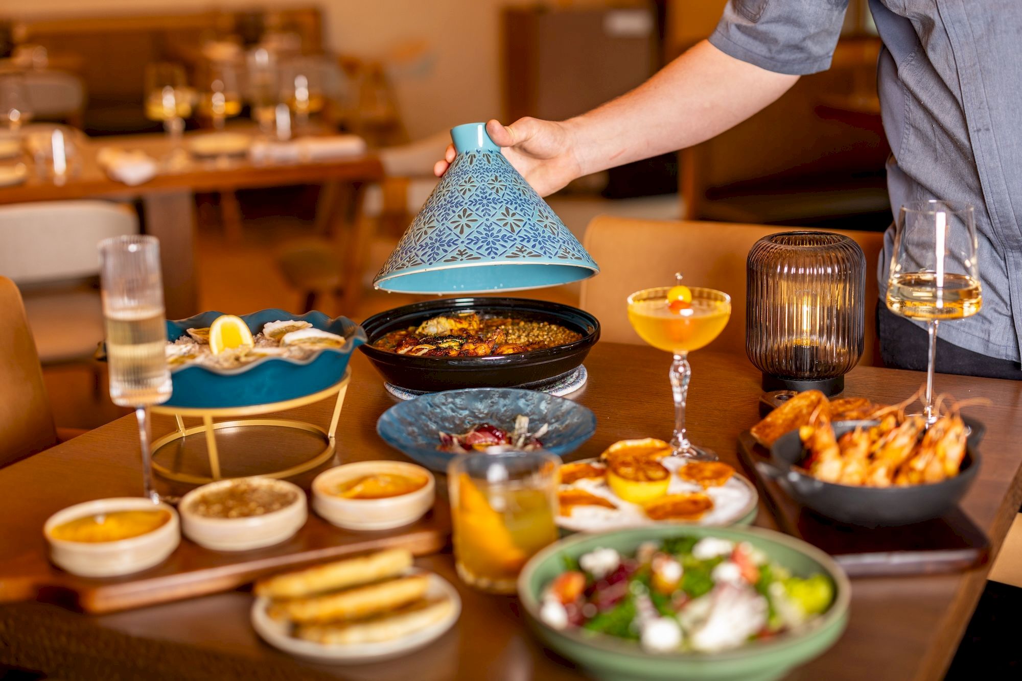 A server lifts a decorative blue cloche over a chafing dish at a catered meal table with drinks, salads, and platters.